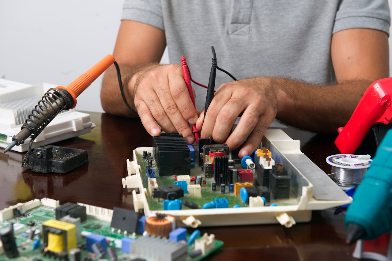 Man working on an electronic circuit board with tools on a table, showing close-up of hands and components.